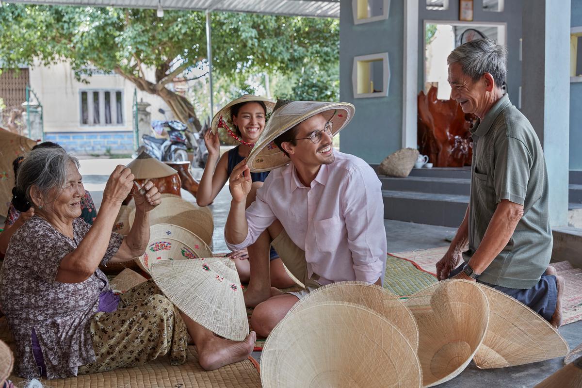 Un couple de voyageurs lors d'un échange souriant et authentique avec une tisseuse de nón lá (chapeaux coniques) au Vietnam. Idéal pour un voyage en couple au Vietnam ou un voyage privé vietnam cambodge axé sur l'expérience.